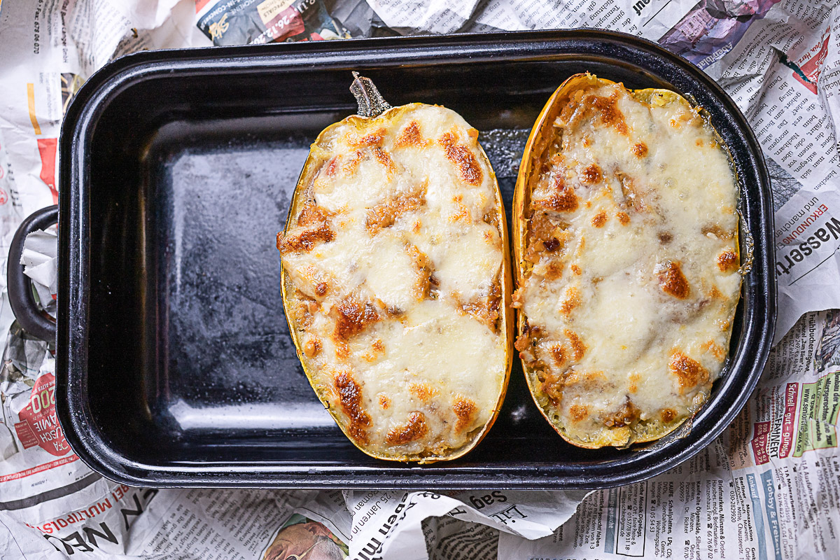 zwei halbe mit roten Linsen, Tomaten, Schafskäse und wenig Hackfleisch gefüllter Spaghettikürbis mit Käsehaube in einer schwarzen Reine
