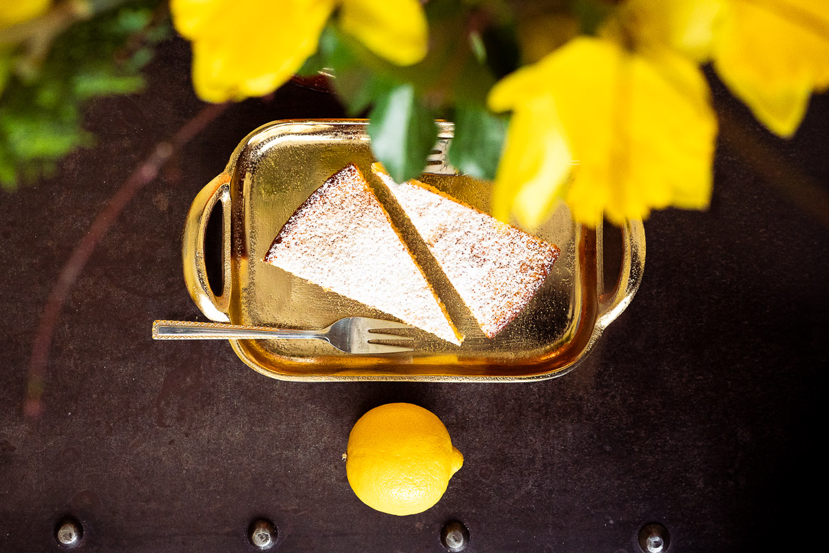 Zwei Stücke mit Puderzucker bestäubter Polentakuchen und eine Kuchengabel auf einem kleinen goldenen Tablett. Daneben liegt und eine Zitrone unter gelben Blumen.