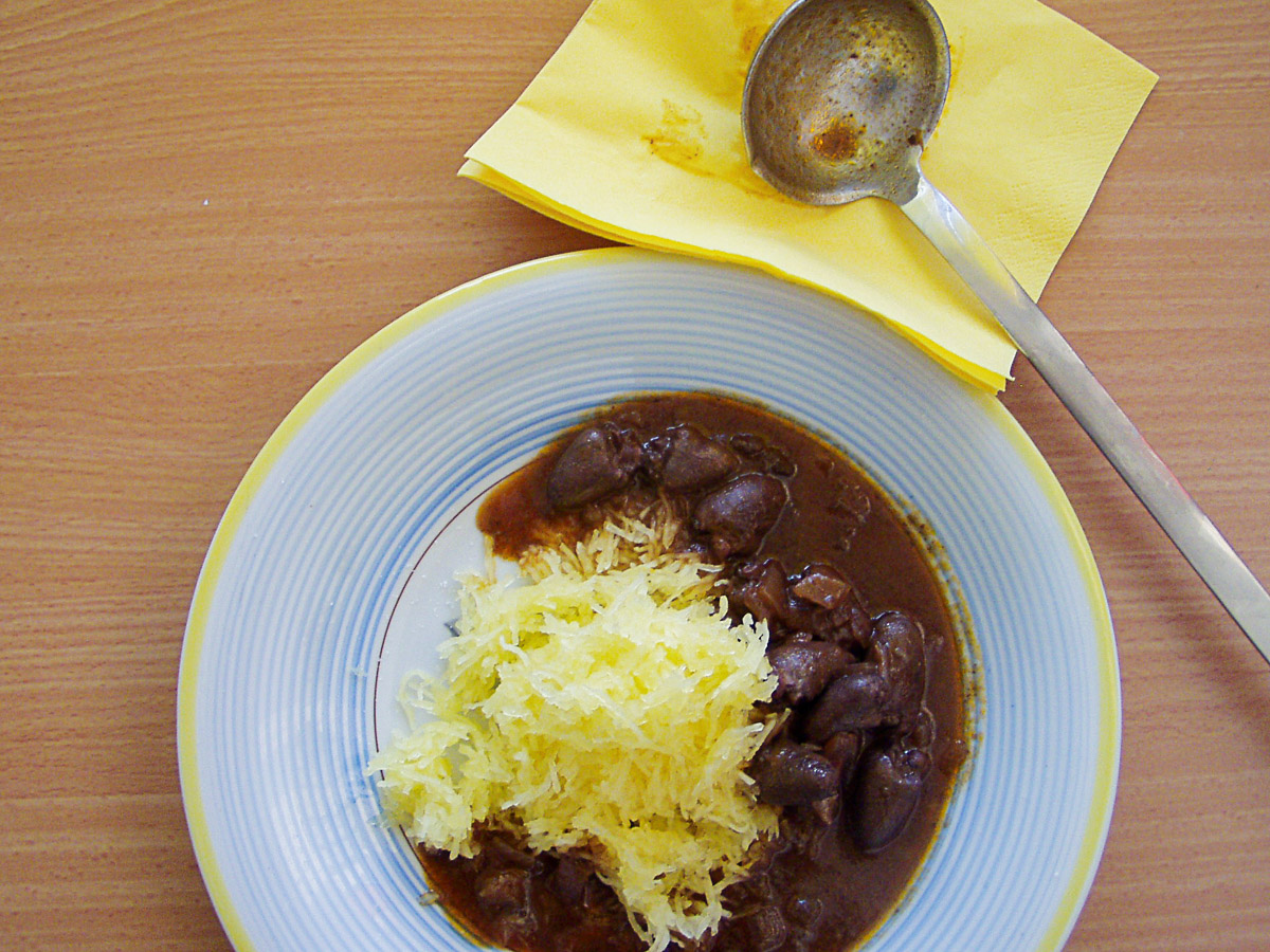 Spaghettikürbis mit Hähnchenherzengulasch auf blau und gelb umrandetem Teller. Daneben liegt eine benutzte Kelle auf gelber Serviette.