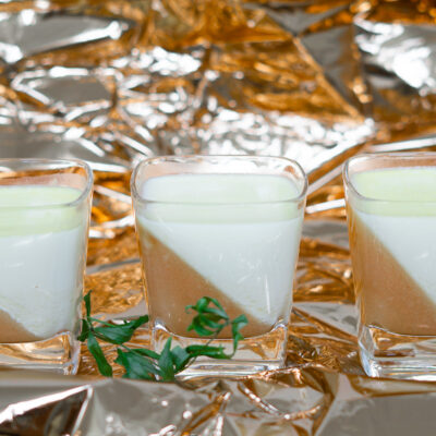 3 Dessertgläser mit Bellini-Waldmeister-Panna Cotta und ein Zweig Waldmeister auf gleichfarbiger Folie wie das Pfirsischgelee im Glas