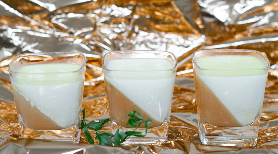 3 Gläser mit Waldmeister-Panna cotta und Pfirsischgelee stehen auf farblich passender Kupferfolie. Davor ein Zweiglein getrockneter Waldmeister