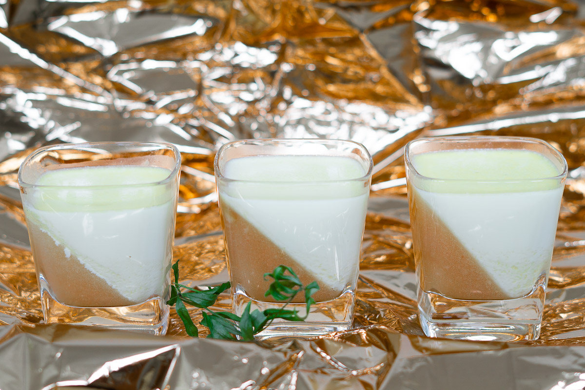 3 Dessertgläser mit Bellini-Waldmeister-Panna Cotta und ein Zweig Waldmeister auf gleichfarbiger Folie wie das Pfirsischgelee im Glas